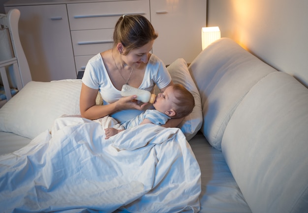 Mulher jovem e bonita dando leite da mamadeira para o bebê à noite