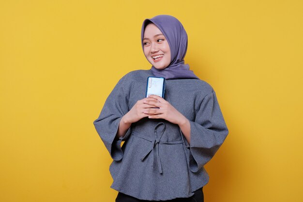 Mulher jovem e bonita de aparência amigável usando um hijab com um lindo sorriso sincero, sentindo-se grata segurando um telefone celular