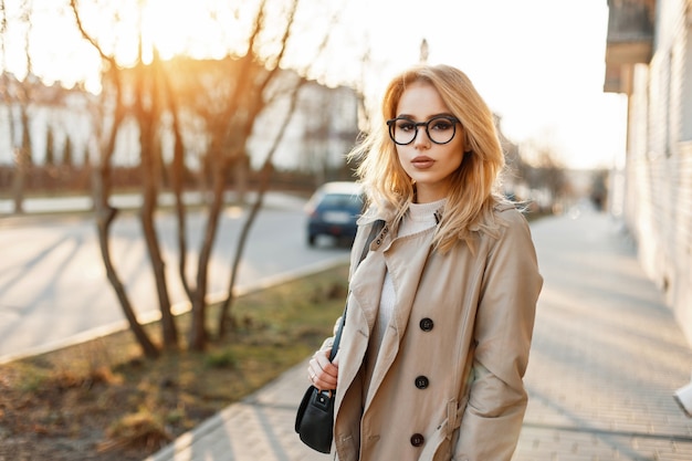 Mulher jovem e bonita de óculos elegantes e um casaco moderno com bolsa no dia de primavera ao pôr do sol