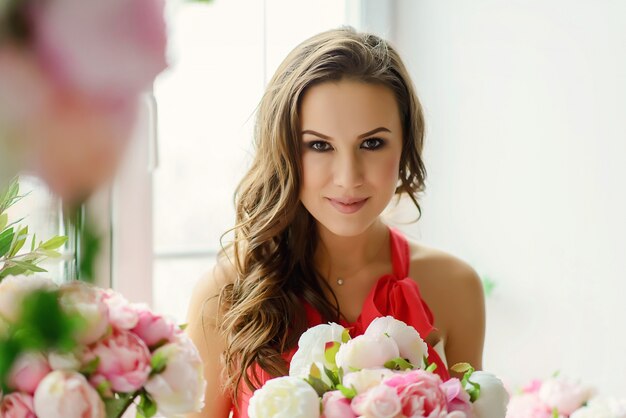 Mulher jovem e bonita de olhos castanhos com um buquê de flores peônias nas mãos perto da janela.