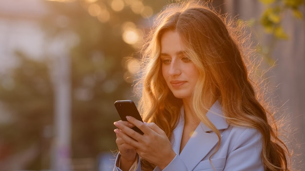 Mulher jovem e bonita de pé na rua ao ar livre em raios de sol ao pôr do sol olhando para o celular respondendo mensagem on-line assistindo vídeo com smartphone conversando desfrutando de internet wifi