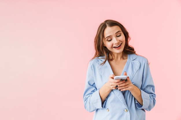 Mulher jovem e bonita de pijama em pé isolado sobre um fundo rosa, usando telefone celular