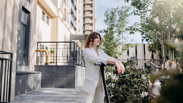 Mulher jovem e bonita de terno branco em pé com sua scooter elétrica perto de um edifício moderno na cidade e desviar o olhar