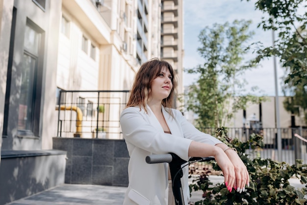 Mulher jovem e bonita de terno branco em pé com sua scooter elétrica perto de um edifício moderno na cidade e desviar o olhar