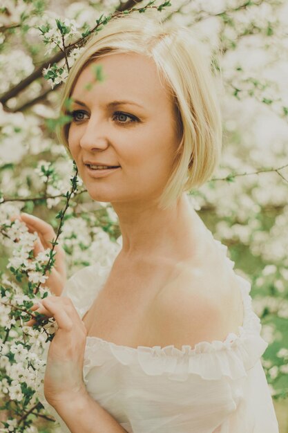 Mulher jovem e bonita de vestido branco com um corte de cabelo loiro curto e ombros nus olha para um galho de cerejeira florescendo no jardim ensolarado na primavera.