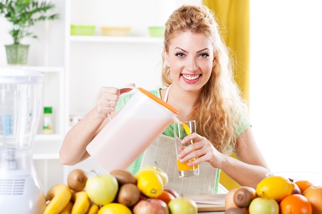 Mulher jovem e bonita derramando suco de laranja no vidro em uma cozinha. olhando para a câmera.