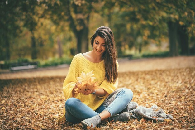 Mulher jovem e bonita desfrutando na floresta ensolarada em cores de outono. ela está segurando folhas amarelas douradas e se divertindo.