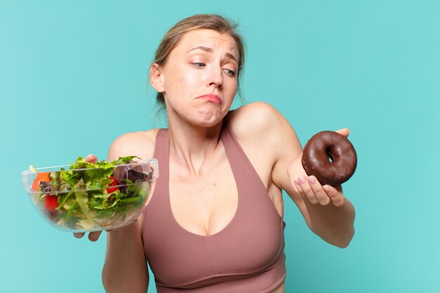 Mulher jovem e bonita do esporte em dúvida ou com uma expressão incerta segurando um sanduíche e um donut