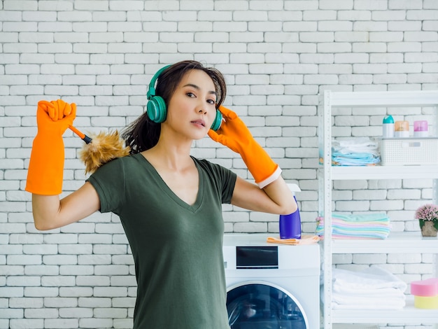 Mulher jovem e bonita, dona de casa usando luvas de borracha laranja, ouvindo música com fone de ouvido verde e segurando o espanador com rosto forte na parede de tijolo branco em casa.