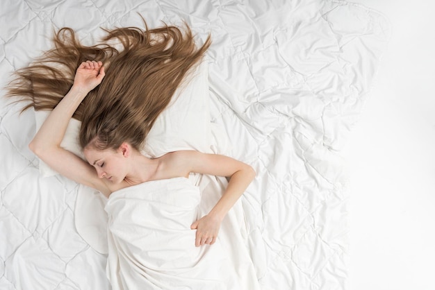 Mulher jovem e bonita dorme em sua cama em lençóis brancos em casa. Retrato de vista superior da menina dormindo com cabelo longo solto. Copie o espaço.