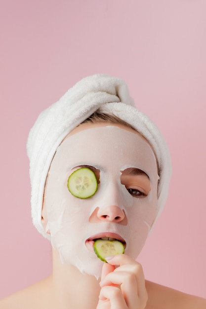 Mulher jovem e bonita é aplicar uma máscara de tecido cosmético em um rosto com pepino