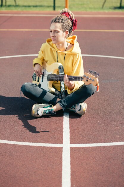Mulher jovem e bonita e elegante tocando guitarra pensativamente enquanto está sentada no campo de esportes