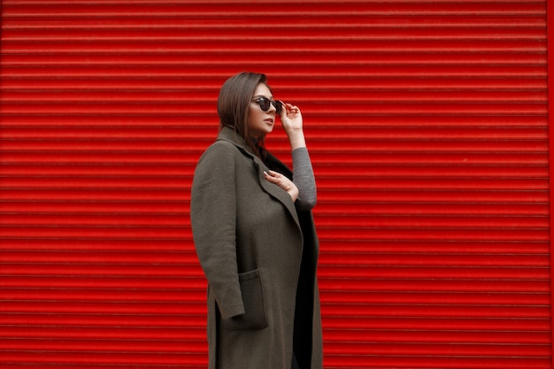 Mulher jovem e bonita elegante com óculos de sol da moda em poses de casaco da moda perto da parede de metal vermelha