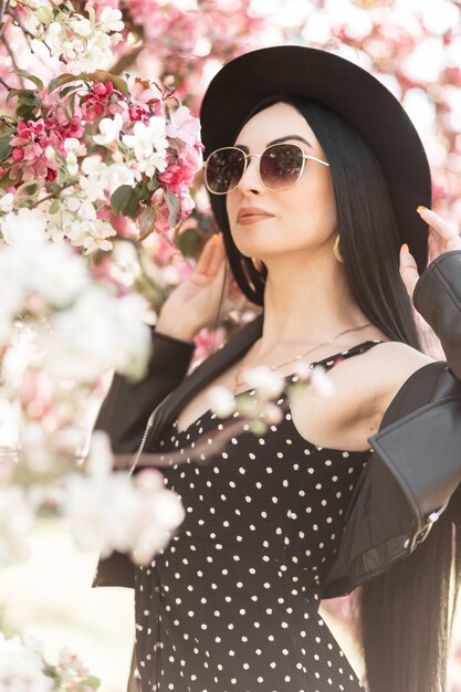 Mulher jovem e bonita elegante com roupas da moda, chapéu e óculos de sol perto de flores na primavera