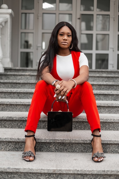 Mulher jovem e bonita elegante negra com a pele limpa em um elegante terno vermelho com bolsa da moda em sapatos sentar nos degraus de pedra na cidade. Modelo de moda garota africana descansar na escada na rua na primavera.