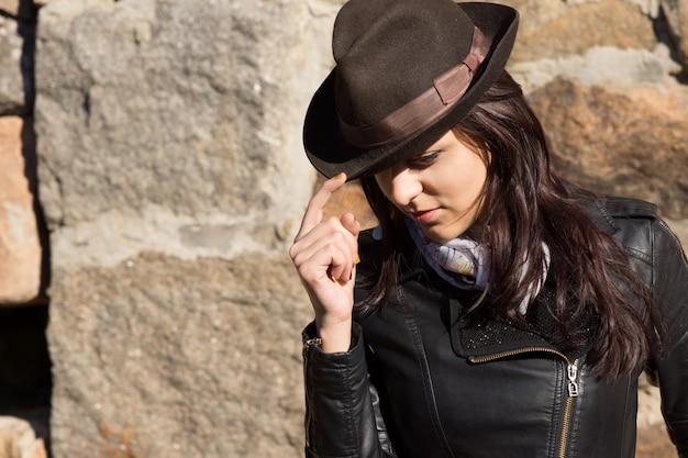 Mulher jovem e bonita elegante usando chapéu fedora marrom e jaqueta de couro preta posando em frente a uma velha parede de pedra com uma expressão facial pensativa legal