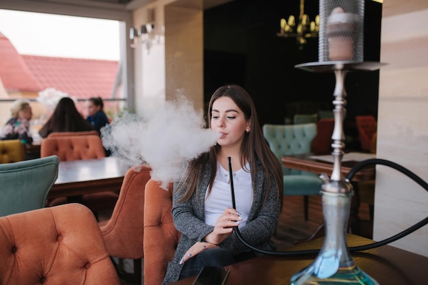 Mulher jovem e bonita em casaco cinza fuma narguilé no terraço do restaurante Smoked