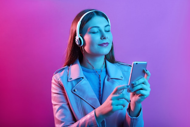 Mulher jovem e bonita em fones de ouvido, ouvindo música com os olhos fechados, isolada sobre o espaço rosa neon, vestindo uma jaqueta de couro, tem cabelo comprido