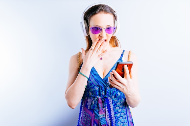 Mulher jovem e bonita em fundo branco usando óculos de verão e fones de ouvido sem fio, olhando para smartphone mostrando surpresa. seu conceito sair de férias. oferecer. anúncio com espaço de cópia