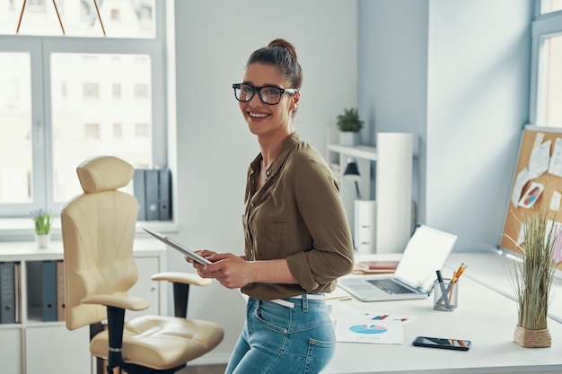 Mulher jovem e bonita em moda casual inteligente sorrindo em pé no escritório