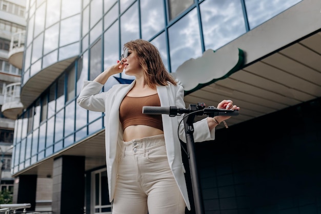 Mulher jovem e bonita em óculos de sol e terno branco em pé com sua scooter elétrica perto de um edifício moderno e desviar o olhar