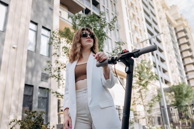 Foto mulher jovem e bonita em óculos de sol e terno branco em pé com sua scooter elétrica perto de um edifício moderno na cidade e desviar o olhar