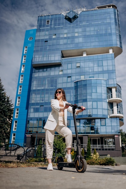 Mulher jovem e bonita em óculos de sol e terno branco em pé em sua scooter elétrica perto de um edifício moderno e desviar o olhar