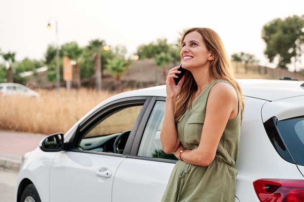 Foto mulher jovem e bonita em pé encostado no carro dela enquanto fala em seu smartphone.