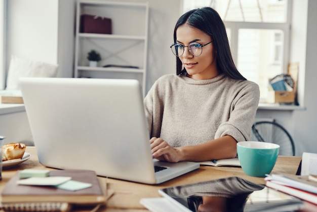 Mulher jovem e bonita em roupas casuais usando laptop e sorrindo enquanto trabalha dentro de casa