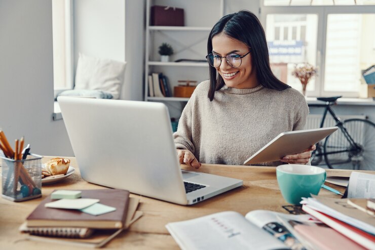 Foto mulher jovem e bonita em roupas casuais usando laptop e sorrindo enquanto trabalhava dentro de casa