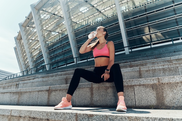 Mulher jovem e bonita em roupas esportivas bebendo água enquanto está sentado ao ar livre