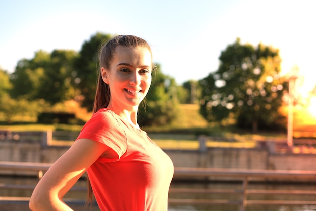 Mulher jovem e bonita em roupas esportivas, sorrindo e olhando uma câmera durante o exercício ao ar livre.