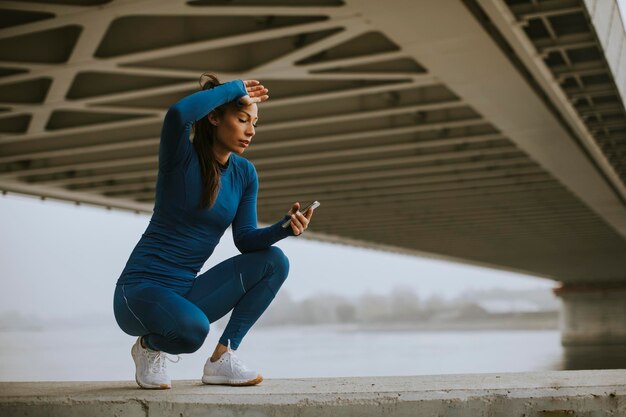 Mulher jovem e bonita em um agasalho de treino azul monitorando o progresso dos exercícios em um aplicativo de condicionamento físico à beira do rio na manhã de outono