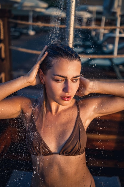 Mulher jovem e bonita em um maiô com uma figura perfeita no chuveiro à luz do sol à beira-mar Garota com bronzeado dourado de férias