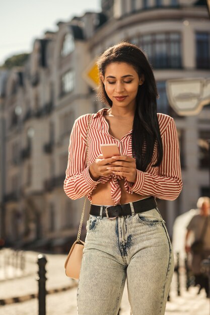 Mulher jovem e bonita em um tempo ensolarado em pé ao ar livre com um smartphone e olhando para a tela