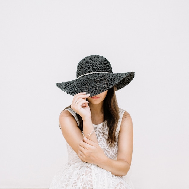 Mulher jovem e bonita em um vestido branco com chapéu preto perto de uma parede branca