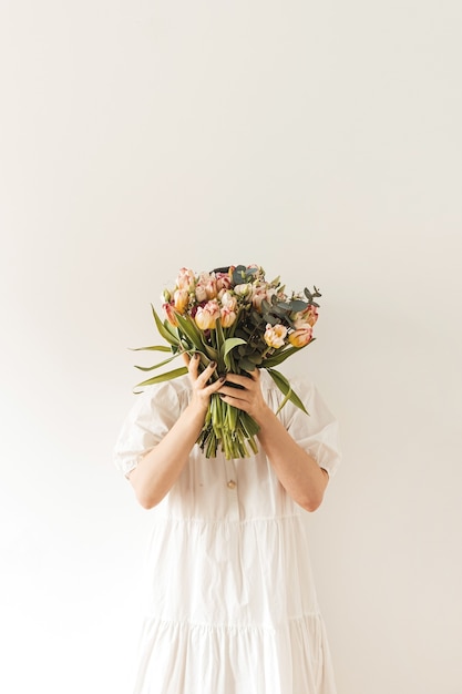 Mulher jovem e bonita em um vestido branco segurando um buquê de flores de tulipa nas mãos contra uma parede branca