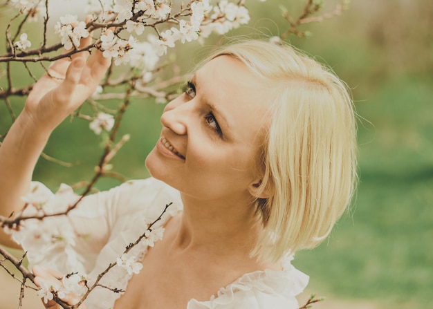 Mulher jovem e bonita em um vestido de chiffon branco com um corte de cabelo loiro curto olha para um galho de cerejeira florescendo, admira e se alegra na primavera.