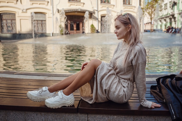 Mulher jovem e bonita em um vestido de malha bege caminha pelas ruas de Moscou, com fontes ensolaradas de clima de primavera nas ruas