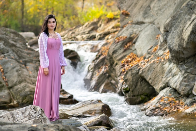 Mulher jovem e bonita em um vestido muito elegante em pé perto do rio da pequena montanha com água em movimento rápido.