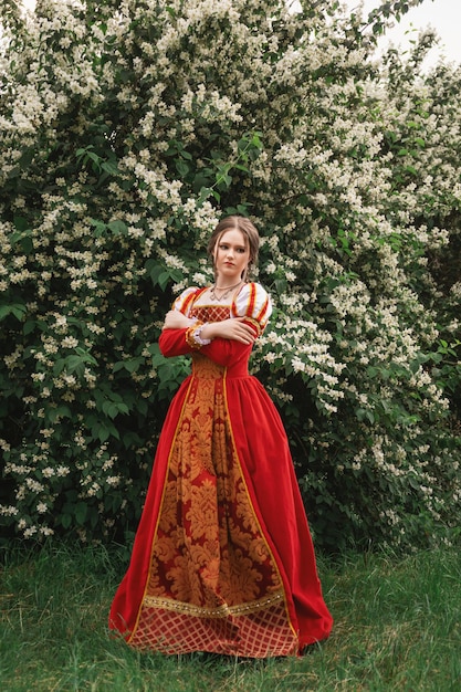 Mulher jovem e bonita em um vestido vermelho medieval está de pé no jardim com flores brancas