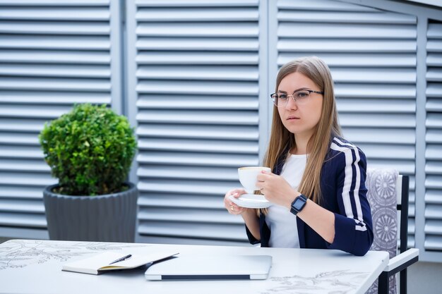 Mulher jovem e bonita em uma camiseta branca está bebendo café e trabalhando em um laptop e sorrindo enquanto está sentado ao ar livre em um café. laptop de mulher jovem para o trabalho. freelancer feminina trabalhando em um laptop