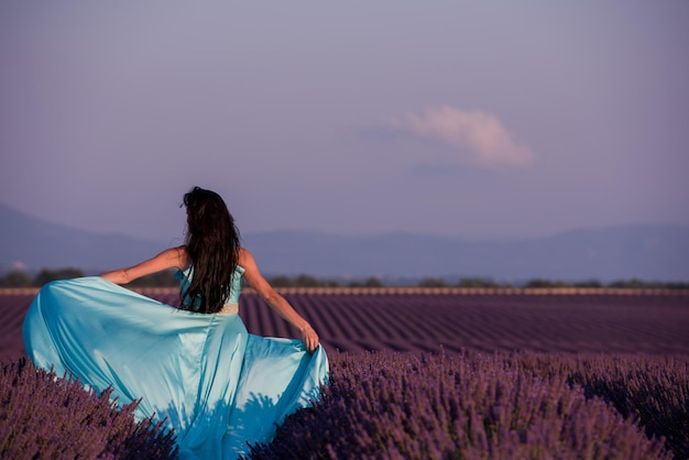 mulher jovem e bonita em vestido ciano relaxando e se divertindo no vento no campo de flores de lavanda roxa