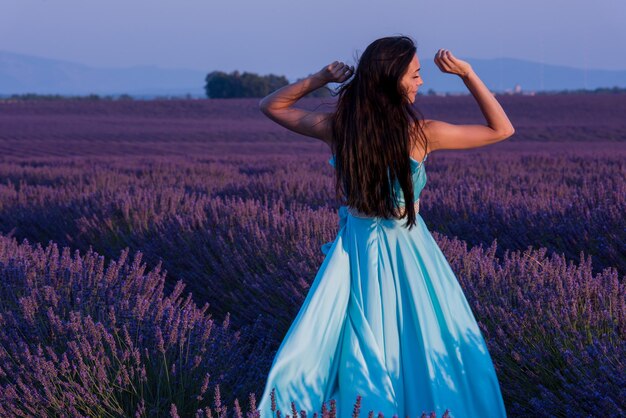 mulher jovem e bonita em vestido ciano relaxando e se divertindo no vento no campo de flores de lavanda roxa