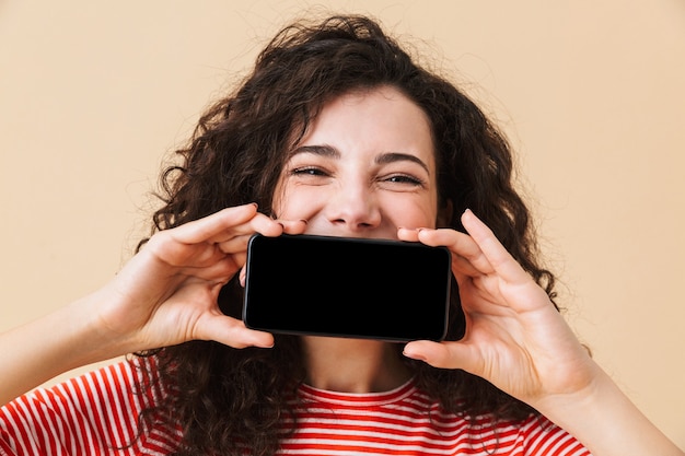 Mulher jovem e bonita encaracolada mostrando o visor pelo telefone celular, cobrindo a boca.