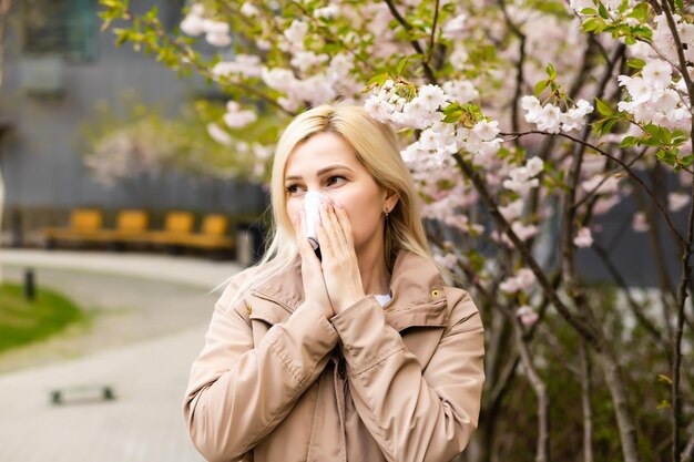 Mulher jovem e bonita espirrando na frente da árvore florescendo. Conceito de alergia de primavera.