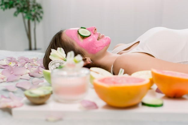 Mulher jovem e bonita está ficando máscara de argila facial no spa, deitado com pepinos nos olhos. Vista lateral.