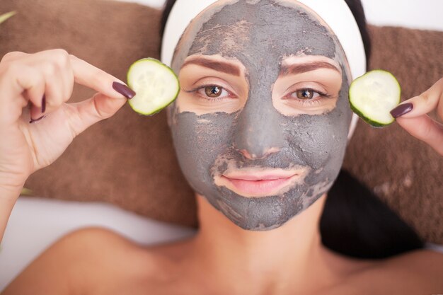 Mulher jovem e bonita está recebendo máscara facial de argila no spa, deitada com pepinos nos olhos