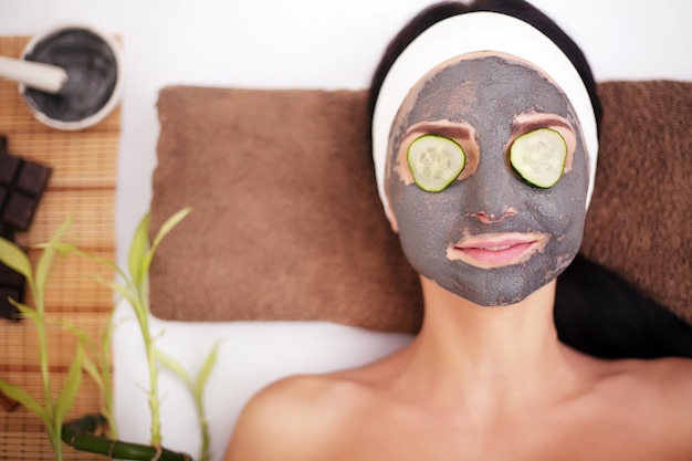 Mulher jovem e bonita está recebendo máscara facial de argila no spa, deitada com pepinos nos olhos