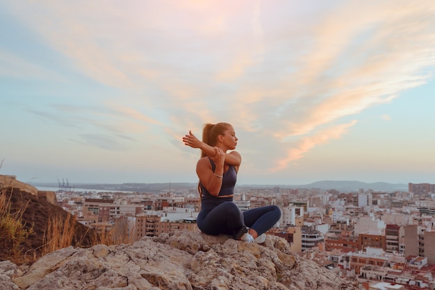 Mulher jovem e bonita faz ioga ao ar livre, no topo de uma montanha na cidade.